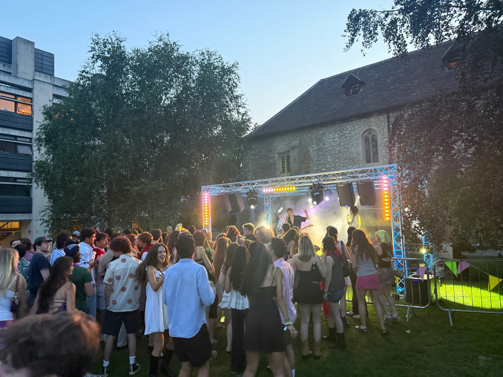 A band performing beneath the illuminated truss at the Cambridge University festival, with an audience gathered on the lawn